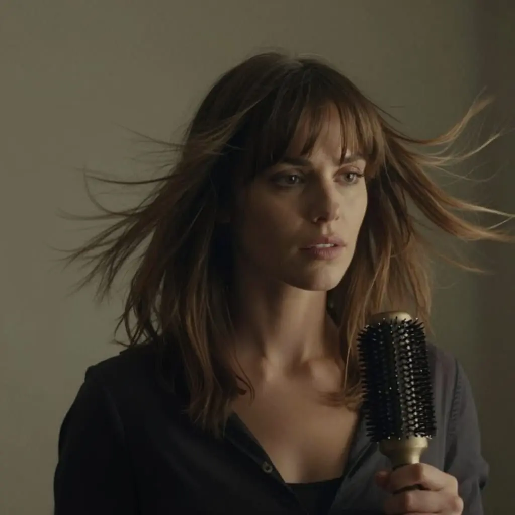 Femme coiffant sa frange rideau avec une brosse ronde, tête légèrement inclinée, cheveux mi-longs balayés par le vent